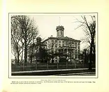 A black and white photo with a yellowing background, showing the original building as it appeared circa 1904.  The building is neoclassical in style, smaller than the current one, with Ionic columns framing the entrance, a tower in the middle, an iron fence encircling it, and trees all around.  There is a caption reading "Indiana Institute for the Blind.  Erected 1847. Is one of the old landmarks of Indianapolis. All of the common school and many of the high school branches are taught the inmates.  They are also given industrial training and instruction in music"