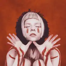 Aurora is against a reddish background with her eyes closed and thumbs under her chin while her palms are open. She has red marks on her skin and a black scarf around her neck.