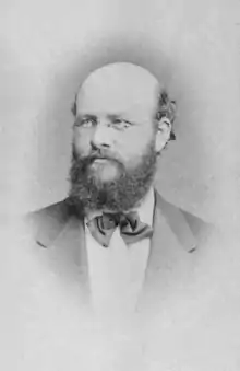 A photograph of a bearded white man with male-pattern baldness wearing glasses