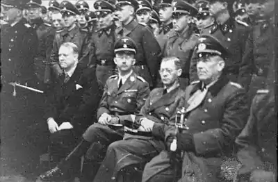 A black and white image of a group of men, most of whom are dressed in the uniforms of military officers. One man, seated towards the front is the only man not in uniform and is dressed in a dark coloured suit.