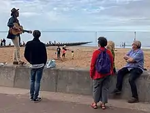 A photograph of a busker playing the guitar to a crowd at Beach Busk 2022