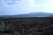 Photograph of a long, flat mountain ridge on the far horizon, with layers to the fore: foothills of the aforementioned mountain; a sliver of a lake; blanket bog and a fence in the very near foreground.