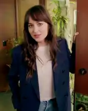 A dark-haired woman wearing a white shirt and a dark blue suit opens the door to her house