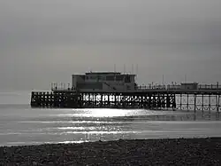 The end of a pier under cloudy skies, illuminated by sunlight reflecting off a calm sea.  There is a small stretch of rocky beach in the foreground.