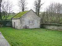 A small, simple chapel seen almost end-on in a grassy burial ground; it is built in stone with a moss-covered roof. On the end is a window with open shutters; on the front face is a door and two shuttered windows.