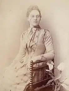 Black and white photograph of a woman, Florence Taylor the astronomer, seated in a chair, wearing a checked close fitting high-necked dress and pince-nez. There is a vase of tulips next to her.