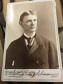 This is a black-and-white photograph of a slender young man with a handwritten signature identifying him as Forrest Robinson at the bottom. The photo captures the man from the chest up with his shoulders facing the front and his head turned slightly to the right against a gray background. He has short, apparently blond or light brown hair parted in the middle and combed back above the ears and light colored eyes. He is clean shaven and has a calm expression with lips closed and eyes directed to the right of the viewer. He is wearing a dark, double-breasted frock coat made of wool with tulip-shaped lapels. A satin or silk lining can be seen on the inner part of the lapel. A crisp, white shirt with a high collar and folded corner wings and a large black tie made of a satin-like material are visible in the short cleavage between the lapels. The coat has visible wrinkles. The photograph is glued to a cardboard backing. The name of the studio, C. M. Galbert, is written in cursive font on the left side and the address of the studio is written on the right side in a smaller cursive font in two lines: illegible number Cherry St., Phila. There is a heraldic emblem centered beneath the photograph. The signature appears in black ink just studio name on the far left with the dedication, Sincerely yours, and the name Forrest Robinson is written in a larger cursive script across the heraldic emblem and the first part of the address. Two thin lines create a frame around the photo and the text.