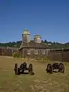 Photograph of the reconstruction of the Fort Ross Chapel, a wooden walled and roofed building with an evergreen-covered hillside behind.