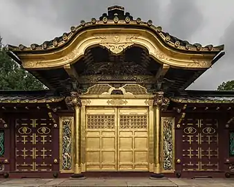 The front of a Shinto Shrine, colored gold and brown, with a golden overhang to the front doors. There are carvings both above and to the sides of the main doors. The sky is overcast and dark.