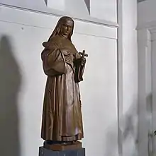 Wooden carving of a full length nun standing holding a large quill pen in her right hand and a crucifix in her left