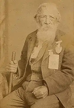 The picture is a sepia toned photograph that shows an African-American man in his eighties wearing a suit adorned with war medals and holding a cane.
