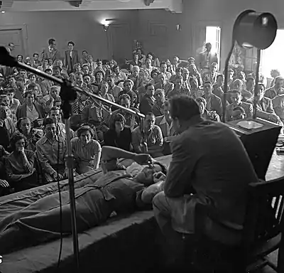 A mostly seated crowd watches as Hubbard, seated on a chair, speaks to a woman lying prone in front of him.
