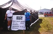 Two people standing outside a large black tent, beside a sign reading 'HRS public health services. Sick call Enfermeria 8am–6pm. Emergencies Emergencia 24 hours.'