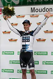 A man in his early twenties wearing a white and black cycling jersey with blue trim and a matching cap. He is holding his arms up in a posture of victory, and holds a bouquet in his right hand.