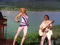 A blonde female dressed in a white leotard with purple stripes stands in front of an image of a lake. To her right, a man dressed in all white plays guitar