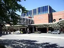 Three-story brick building with blue windows; a glass pedestrian bridge travels from building to unseen parking garage on left, crossing entrance road. "ALBANY INTERNATIONAL AIRPORT" sign is visible on side.