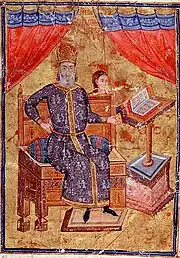 Elderly bearded man sitting on a wooden throne, pointing to a lectern with a book open on it. He is dressed in a long blue white-embroidered and gold-edged kaftan, and wears a domed golden-red hat.