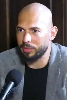 A bald man with a beard looks at something off-camera with a microphone near his face.