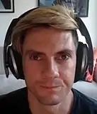 A man with blond hair and headphones smiling at the camera.