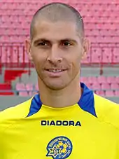 A upper-body photograph of bald white man, wearing a yellow association football shirt.