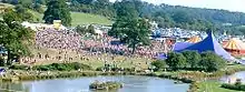 An aerial shot of a large gathering of people in a grassy area beside a lake. Several large tents are visible.