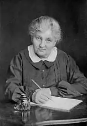 A grandmotherly woman of about 70 is seated, pen in hand, at a desk with a writing pad.