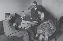 Black-and-white photograph of three men in suits and one woman seated in a room and looking at an open newspaper