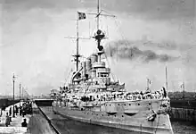 A large warship passes through a narrow canal while numerous onlookers watch from the edge of the canal.