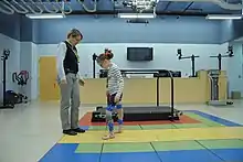 A girl wearing leg braces walks towards a woman in a gym, with a treadmill visible in the background.