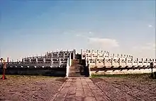 Color photograph of a three-level stone structure with railings on each level, viewed from the outside, facing a staircase that leads to the top level.