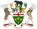 A central shield with the upper part showing the red cross of St. George and the lower part showing three golden maple leaves on a green background. There is a black bear on top of a knight's helmet above the shield with a moose to the left and a Canadian deer to the right. The province's motto "Ut incepit Fidelis sic permanent", Latin for "Loyal she began, loyal she remains" is written below the crest.