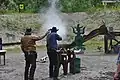 A Cowboy action shooter firing a lever action rifle at steel targets. The Range Officer to the left is holding a shooting timer to measure the time.