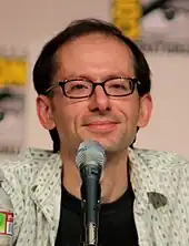 A closeup of a man in front of a microphone. He has a receding hairline and wears dark-framed glasses.