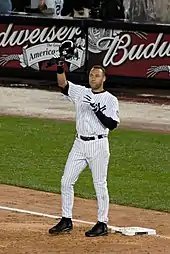 A man in a white baseball uniform with navy pinstripes removes his helmet to salute the crowd, which is cheering for him.