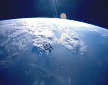 A cluster of modules and feathery solar arrays floats in the middle distance before an image of the Earth and the blackness of space above its horizon. Sunrays project from the top centre of the image.