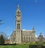 A tall clock tower in shape rather like Big Ben at Westminster; behind this is a Gothic chapel with an apse on the left and a turret on the right