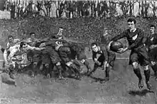 Painting of the aftermath of a rugby scrum, with a player from one team running with the ball towards the opposition goal-line while an opponent runs to intercept him.