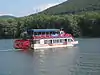 A red, white and blue paddlewheel boat with a sign "Hiawatha" on a river with a forested mountain behind