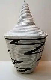 Photograph depicting a bowl shaped off-white woven basket with tall conical lid and black zigzag pattern
