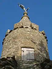 Circular tower with wooden door. On the top is a small statue.