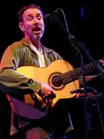 A man with a goatee singing into a microphone playing a guitar.