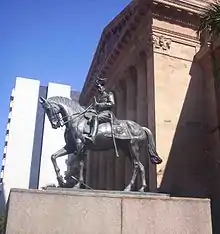 Equestrian statue in dark grey metal of George V in military dress uniform on a plinth of red granite outside a Classical building of red sandstone