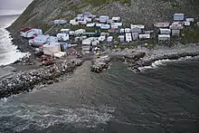 A village of 40–50 mostly small buildings built on a steep, rocky coastline; some structures higher on the hill are built on stilts.