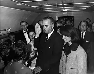 Lyndon B. Johnson raises his hand above an outstretched Bible as his is sworn in as President as Air Force One prepares to depart Love Field in Dallas. Jacqueline Kennedy, still in her blood-spattered clothes (not visible), looks on.