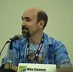 A Caucasian bald man with a black beard looks to the left while speaking into a microphone. A plaque with the Comic-Con logo on the table reads "Mike Stemmle".