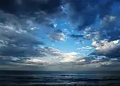 Late daytime view looking far out over an ocean from a beach, which is out of view off the bottom margin. Three-fourths of the shot features a sky marked by heavy cloud cover, which is parting near the middle, revealing a dazzlingly bright cerulean blue sky that darkens near the margins. The ocean is striated with waves coming in parallel to the horizon.