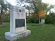 An angled view of a large stone monument with a placard and two nearby white historical markers.