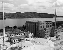 Large building shaped like a cube. In front is a smaller, barn-like building. They are flanked by two smokestacks. There is a river in the background.