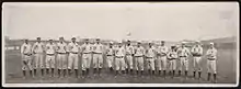 A single row of men in white baseball uniforms with high socks and white baseball caps standing on a baseball field; their uniforms read "NY" across the chest.