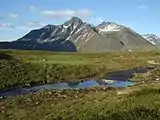 Mountain with two main peaks, composed of dark rock with traces of snow on the sides, against a bright blue sky. In the foreground, a stream cuts through grassland.